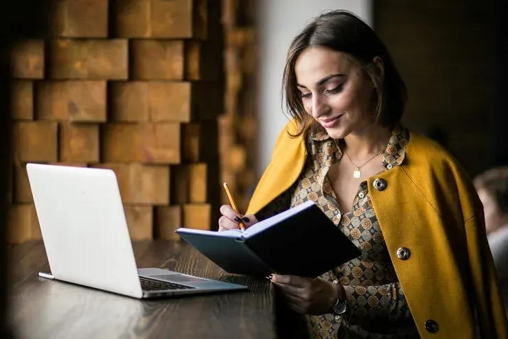 Smiling woman writing in a notebook beside an open laptop
