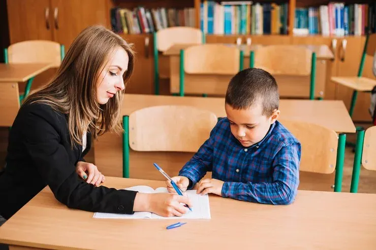 Teacher helping a young boy write at a classroom desk
