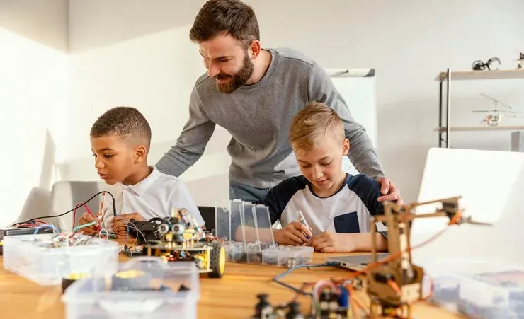 Teacher helping two boys build electronic robots at a workbench